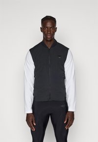 Schwarze ärmellose Weste mit Reißverschluss vorne und Seitentaschen, getragen über einem weißen Langarmshirt, kombiniert mit schwarzen Sporthosen.