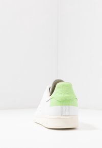 Vita sneakers med en ljusgrön hälflik och präglad logotyp, fotograferade från baksidan på en vit yta och bakgrund.