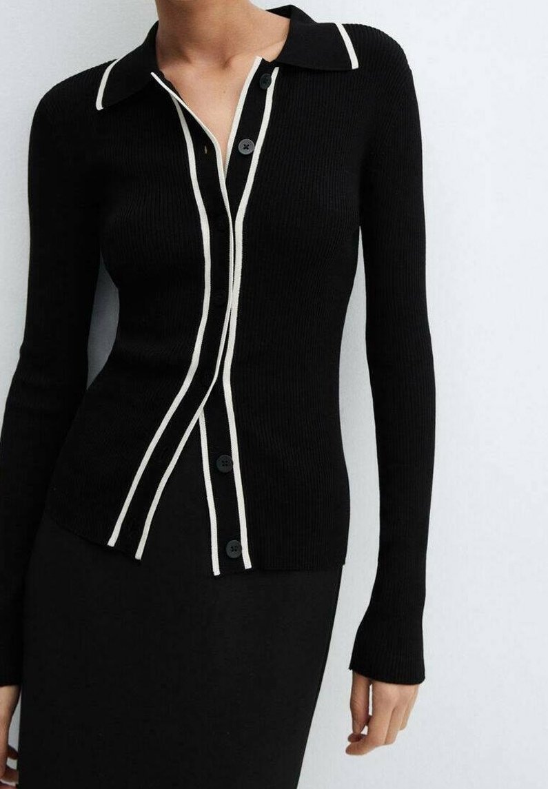 Cardigan noir à manches longues en côtes avec une bordure blanche le long du col et de la patte de boutonnage, porté avec une jupe noire assortie.