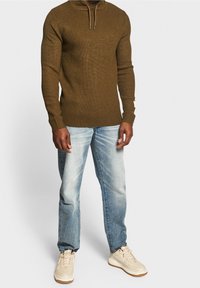 Man met een bruine gebreide hoodie, lichtblauwe verweerde spijkerbroek en beige sneakers staand tegen een witte achtergrond.