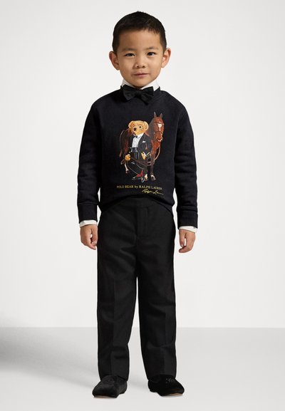 Svart genser med et grafisk motiv av en bjørn i smoking, kombinert med svarte bukser og svarte sko, med en hvit skjorte og sløyfe under.