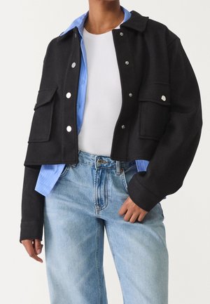 Personne portant une tenue à plusieurs couches avec une veste courte noire, une chemise bleue boutonnée, un haut blanc et un jean bleu clair.