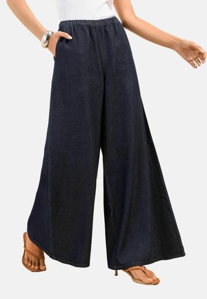 Femme portant un pantalon large noir à taille élastique, un haut blanc sans manches, un bracelet argenté et des sandales marron à bouts ouverts.