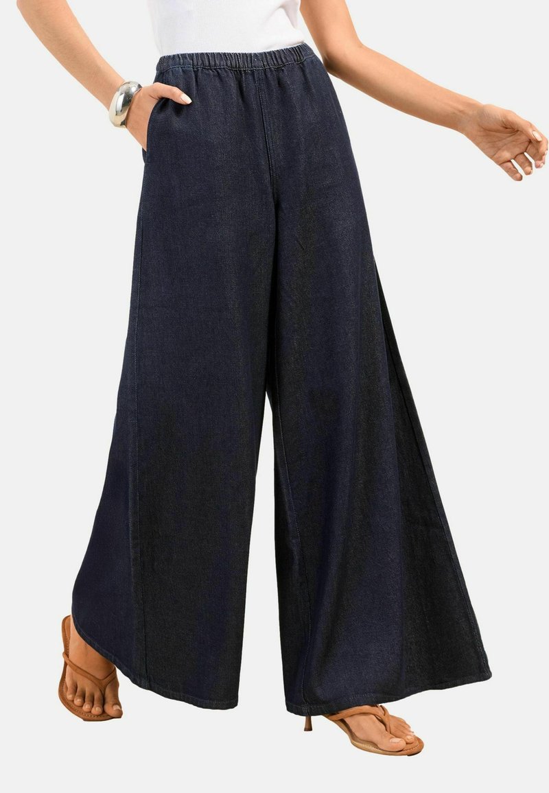 Nő sötét, széles szárú nadrágban, rugalmas derékpánttal, fehér ujjatlan felsőben, ezüst karkötővel és barna nyitott orrú szandálban.