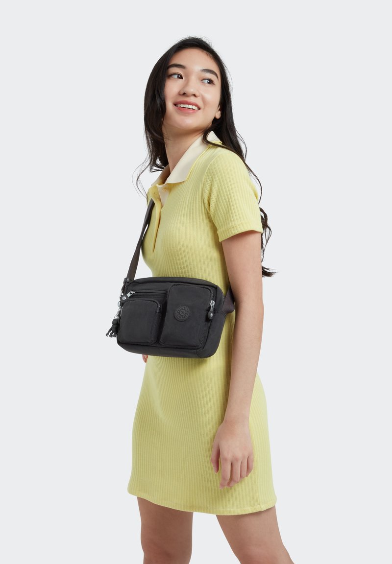 Borsa a tracolla in tessuto nero con due tasche frontali con zip e dettaglio del logo, indossata su un abito giallo chiaro a coste con colletto.