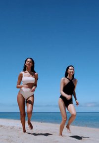 To kvinner løper på en sandstrand. Den ene har på seg en lys rosa bikini, mens den andre har på en svart heldrakt med kryssdesign. Klar blå himmel.