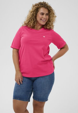 Frau mit lockigem Haar lächelnd, trägt ein hellrosa kurzärmliges Hemd mit kleinem Herzlogo und knielangen Jeansshorts.