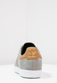 Grå mockaskor med en tan läderhäl, med en guldig Adidas-logotyp och vit gummisula. Bakifrån framhävs de texturerade materialen.