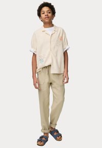 Chemise en lin beige clair avec des motifs brodés colorés, associée à un pantalon kaki clair à coupe décontractée et des sandales bleues avec des boucles.