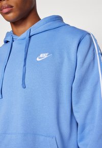 Gaiši zils Nike sudraba ar priekšējo kabatu, kapuci ar aukliņām un balto logotipu uz krūts. Mīksta auduma ar gludu tekstūru un smalku apdari.