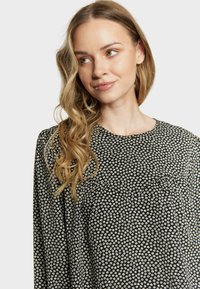 Blouse en tissu noir avec un motif floral blanc, encolure ronde et détails plissés, dotée de manches longues et d'une texture douce.