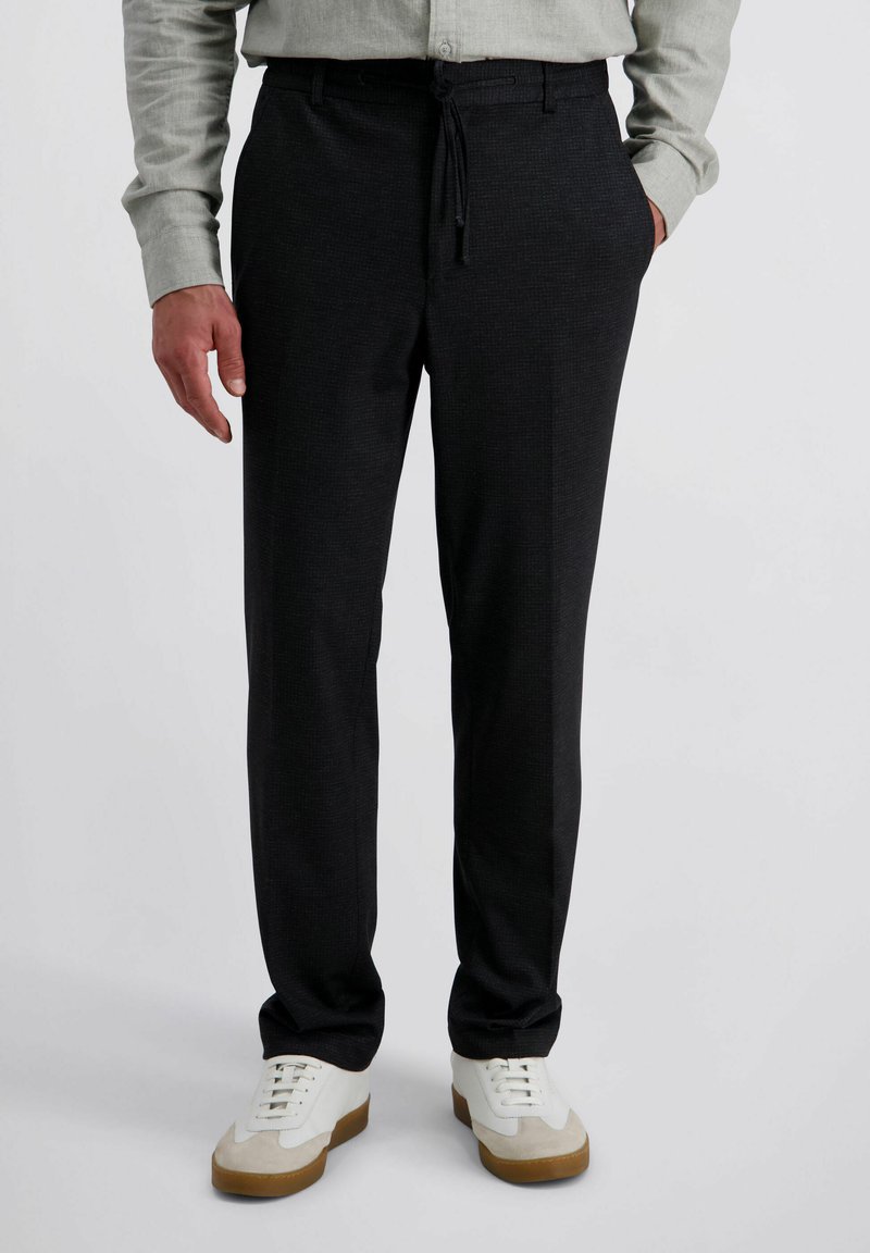 Man in zwarte drawstringbroek, lichtgrijs button-up shirt en witte sneakers met beige accenten.
