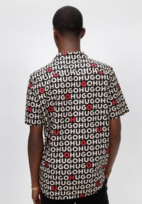 Camicia a maniche corte con un audace motivo testuale ripetuto in nero, rosso e beige. Realizzata in un tessuto leggero con una vestibilità comoda.