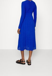 Robe en tricot bleue à manches longues, avec des motifs verticaux et un corsage ajusté. Portée avec des sandales noires.