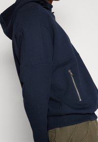 Felpa blu navy con texture a maglia, dotata di tasca laterale con zip e cappuccio con coulisse. Il design presenta cuciture sovrapposte.