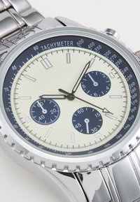 Montre argentée avec bracelet en métal, cadran crème, sous-cadrans bleus et échelle tachymétrique. Présente des marqueurs d'heure élégants et des aiguilles fines.