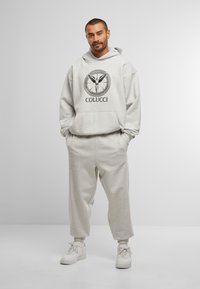 Muž s krátkymi vlasmi a bradou, oblečený v svetlosivej kapucňovej mikine Colucci, zodpovedajúcich teplákových nohaviciach a bielych teniskách, stojí s rukami v vreckách.