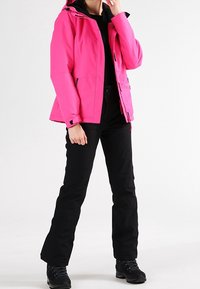 Žena stojící v jasně růžové bundě s kapucí, černých kalhotách a černých turistických botách na bílém pozadí.