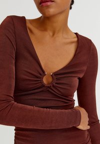 Braunes Langarmoberteil mit tiefem V-Ausschnitt, das ein Ringdetail an der Brust und gerafftes Material für Textur aufweist. Weiches, figurnahes Design.