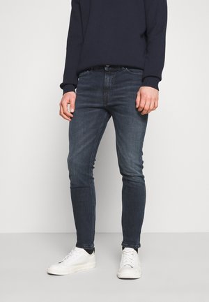 Vaqueros de mezclilla azul oscuro con corte ajustado, ligera decoloración y diseño estructurado. Combinados con zapatillas blancas y un suéter azul marino oscuro.