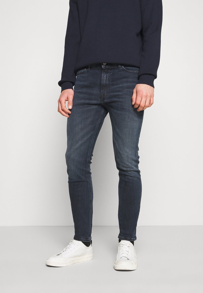Mörkblå denimjeans med slim fit, lätt blekning och strukturerad design. Kombinerat med vita sneakers och en mörkblå tröja.