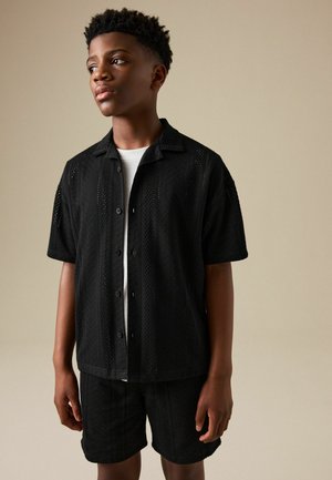 Young man looking to the side wearing a black short-sleeve button-up shirt and matching shorts with a white undershirt.