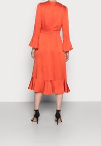 Robe orange à manches longues avec des poignets évasés, une ceinture à la taille et un ourlet froncé. Assortie à des talons aiguilles noirs à bout ouvert.