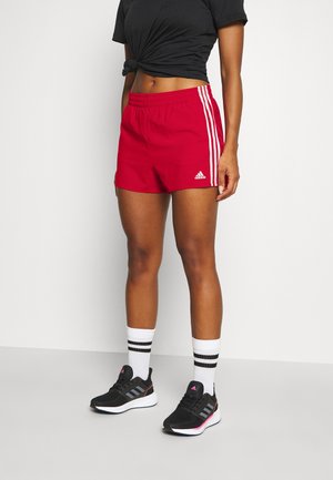 Persoon die rode Adidas-shorts draagt, een zwart geknoopt cropped topje, witte sokken met zwarte strepen en zwarte Adidas-sneakers, staand tegen een effen achtergrond.