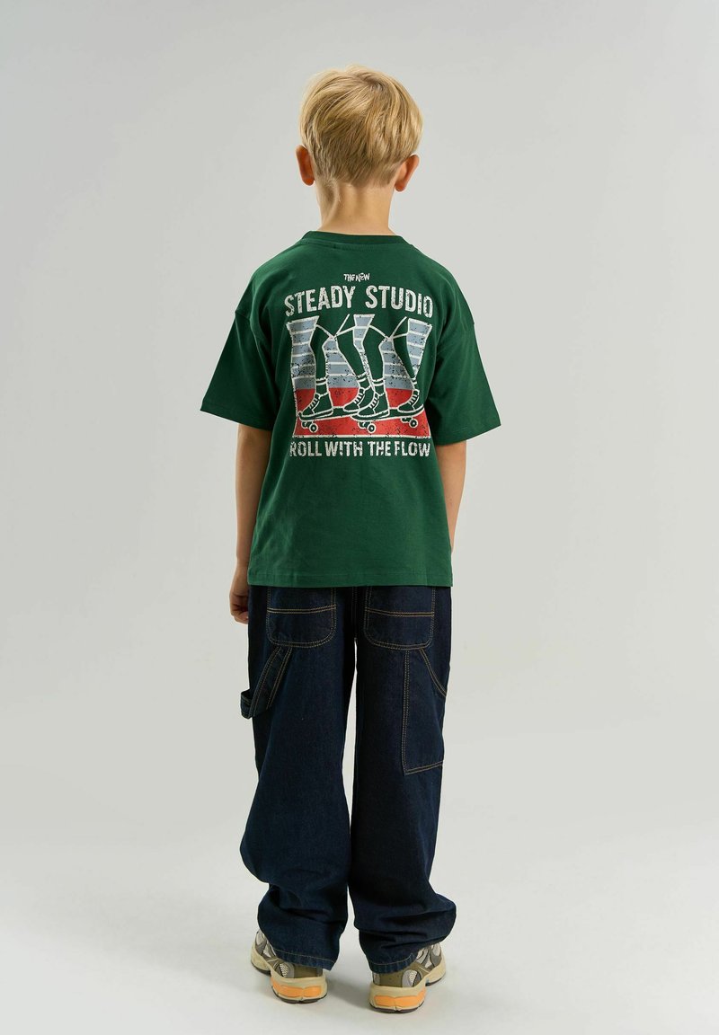 Enfant blond de dos, portant un t-shirt vert avec un motif de skateboard et un texte, un pantalon ample sombre et des baskets beiges sur fond uni.
