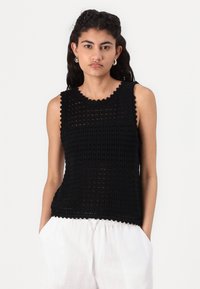 Top sin mangas de ganchillo negro con un escote ondulado, que presenta una trama abierta y un patrón texturizado, combinado con pantalones ligeros y holgados.