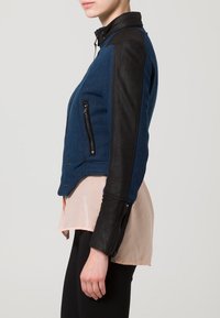 Blå ullblandad jacka med svarta läderslouvar, sidofickor med dragkedja och krage, med en figursydd design och kontrasterande texturer.