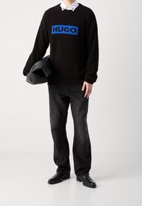 Schwarzer gerippter Pullover mit blauem "HUGO"-Logo, kombiniert mit schwarzen Jeans und schwarzen Schuhen. Das Modell trägt eine schwarze gepolsterte Tasche.