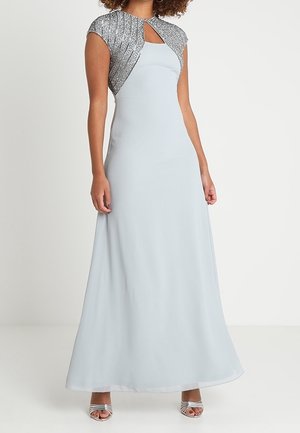 Frau trägt ein hellblaues, bodenlanges Kleid mit silbernem, paillettenbesetztem Bolero und silbernen offenen High Heels, steht vor einem einfarbigen Hintergrund.