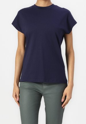 Kobieta ubrana w gładką granatową koszulkę z krótkim rękawem i okrągłym dekoltem oraz dopasowane szare spodnie, stojąca na białym tle.