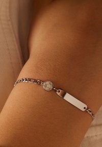 Zilveren schak bracelet met een glad rechthoekig label en een ronde bedel die lijkt op een voetbal, gezet tegen een huidkleurige achtergrond.