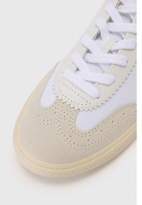 Sneaker med en hvit lerretsoverdel, kremfarget semsket tå, sikksakk- søm langs kanten og hvite snører, med en teksturert gummisåle.