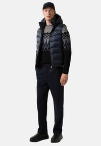 Mann mit schwarzer Mütze, marineblauem Steppwesten, gemustertem grau-schwarzem Pullover, marineblauen Hosen und schwarzen Schuhen, der mit den Händen in den Taschen steht.
