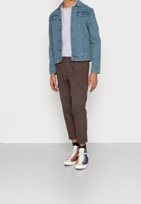 Kurtka dżinsowa w jasnoniebieskim kolorze, biała koszulka, brązowe spodnie typu cargo oraz kolorowe trampki za kostkę z kontrastującymi akcentami i teksturami.