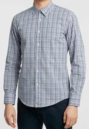 Homme portant une chemise à manches longues boutonnée avec un motif à carreaux verts, bleus et blancs et une poche poitrine à l'avant.
