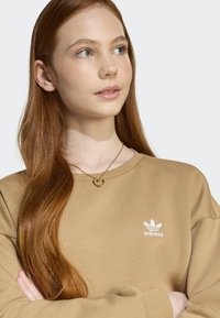 Beige sweatshirt med rund halsringning, med en vit Adidas-logga och mjuk textur. Ett guldhalsband med hjärta kompletterar looken.