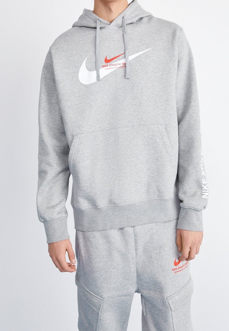 Šedá mikina s kapucí vyrobená z měkkého materiálu, s přední kapsou, stahovací kapucí a logy Nike v červené a bílé barvě na hrudi a rukávu.