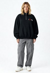 Sweat-shirt en polaire noire avec col montant, fermetures éclair blanches et texte brodé « I ❤️ You » en rouge et blanc. Associé à un pantalon cargo gris et des baskets blanches.