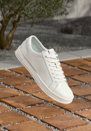 White Calvin Klein leather sneaker with laces resting above wooden square tiles and small stones outdoors by a tree.