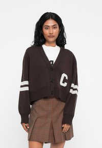 Cardigan castanho cropped com decote em V, quatro botões pretos e riscas brancas nas mangas. Apresenta a letra "C" na parte da frente. Textura suave.