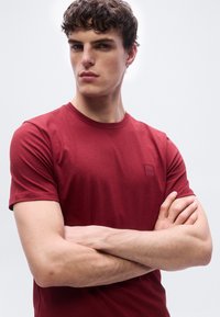 Νεαρός με σγουρά μαλλιά που φορά κόκκινο κοντομάνικο πουκάμισο, στέκεται με τα χέρια σταυρωμένα μπροστά σε απλό φωτεινό φόντο.