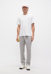 Νέος άνδρας με λευκό καπέλο και μπλουζάκι, γκρι φόρμα, κρατώντας μια μπάλα του τένις, στέκεται μπροστά σε απλό λευκό φόντο.