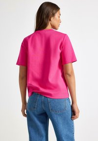 Kvinde med langt brunt hår iført en lys pink kortærmet skjorte og blå højtaljede jeans, der står med ryggen til mod en hvid baggrund.