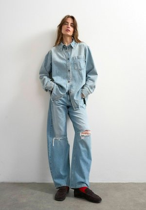 Kvinde står op ad en hvid væg iført en stor lys denimskjorte, revnede vide jeans, røde sokker og mørke loafers.