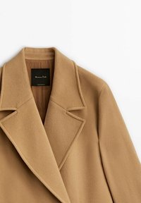 Manteau en laine couleur chameau avec larges revers, étiquette intérieure visible et texture lisse, présenté à plat sur fond blanc.