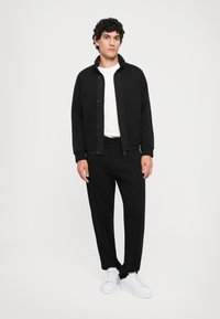 Zwarte zip-up jas met ribgebreide manchetten, gecombineerd met zwarte toegespitste broek en witte sneakers. Effen wit t-shirt eronder, casual fit.